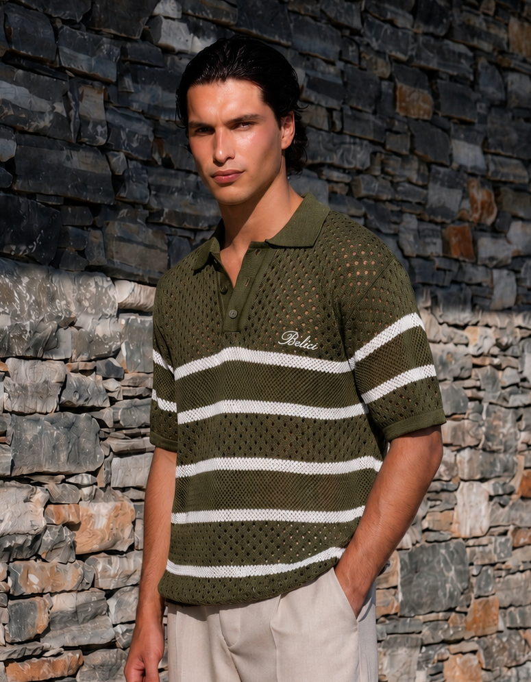 A man wearing a short-sleeve knit polo shirt in olive green with white horizontal stripes, standing against a rustic stone wall. The shirt features a textured open-knit pattern and a small white script logo on the left chest.
